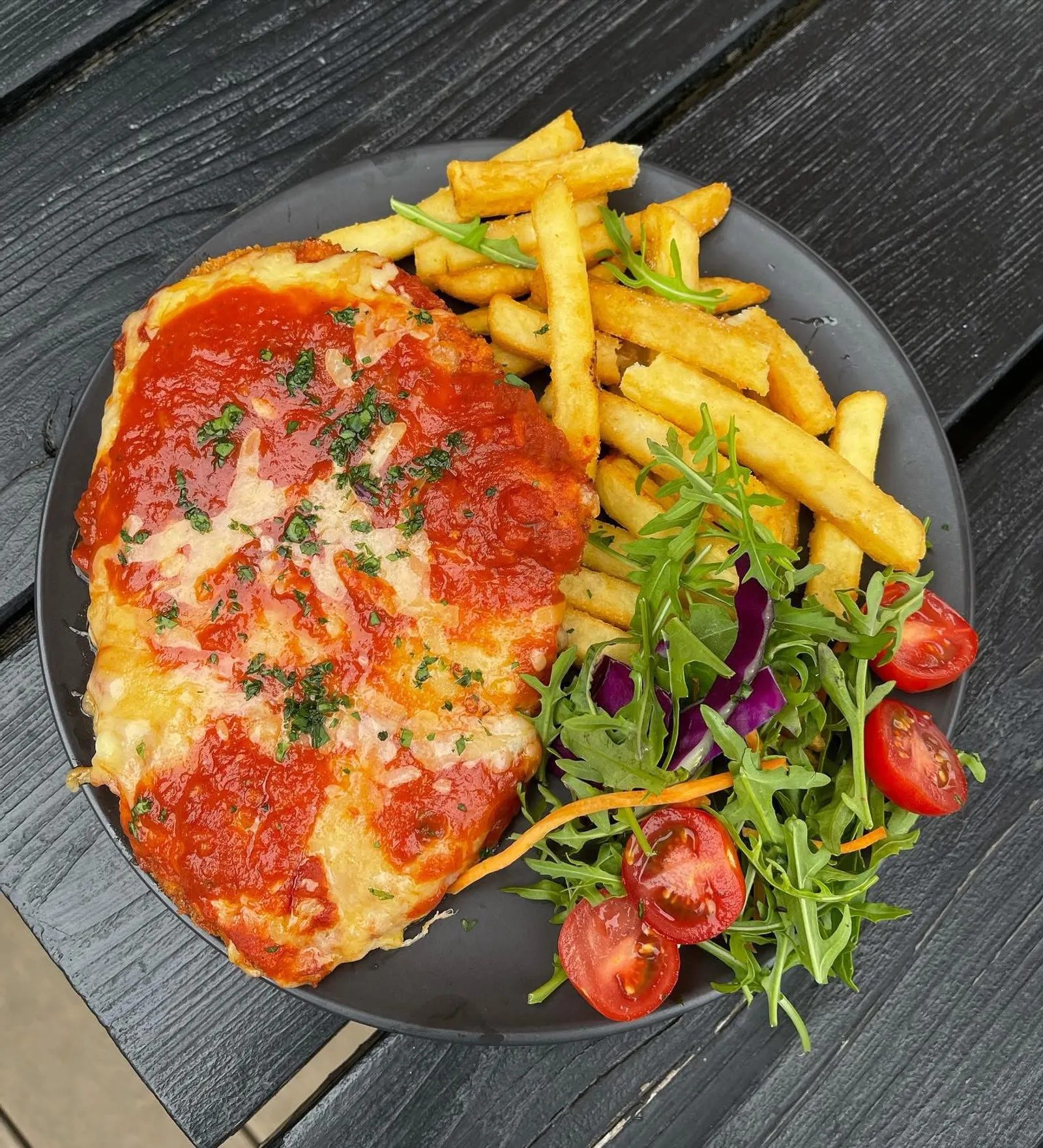 Chicken parmigiana with chips and salad on a dark plate, viewed from above
