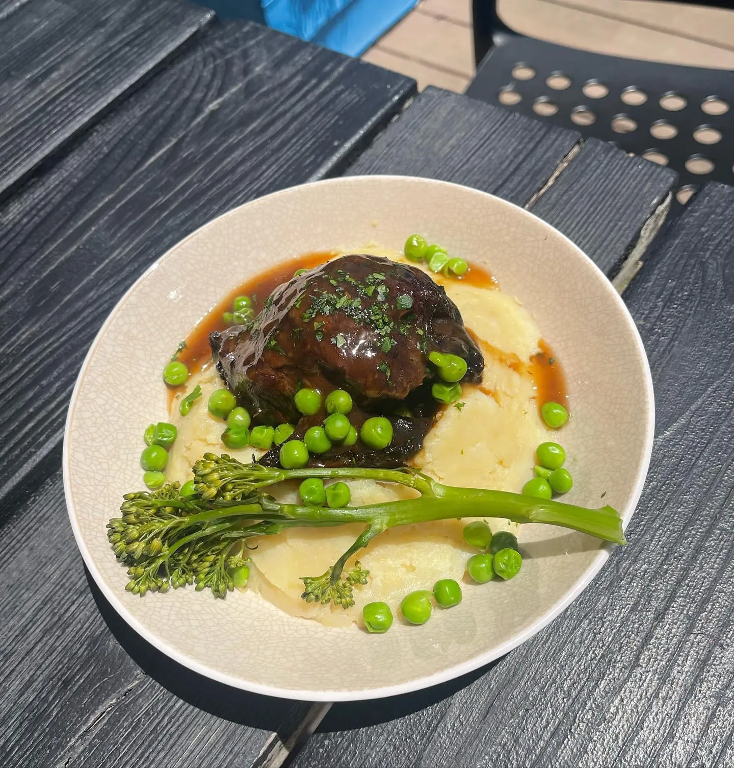 Braised beef cheek on mashed potato with green peas, broccolini, and jus in a white bowl