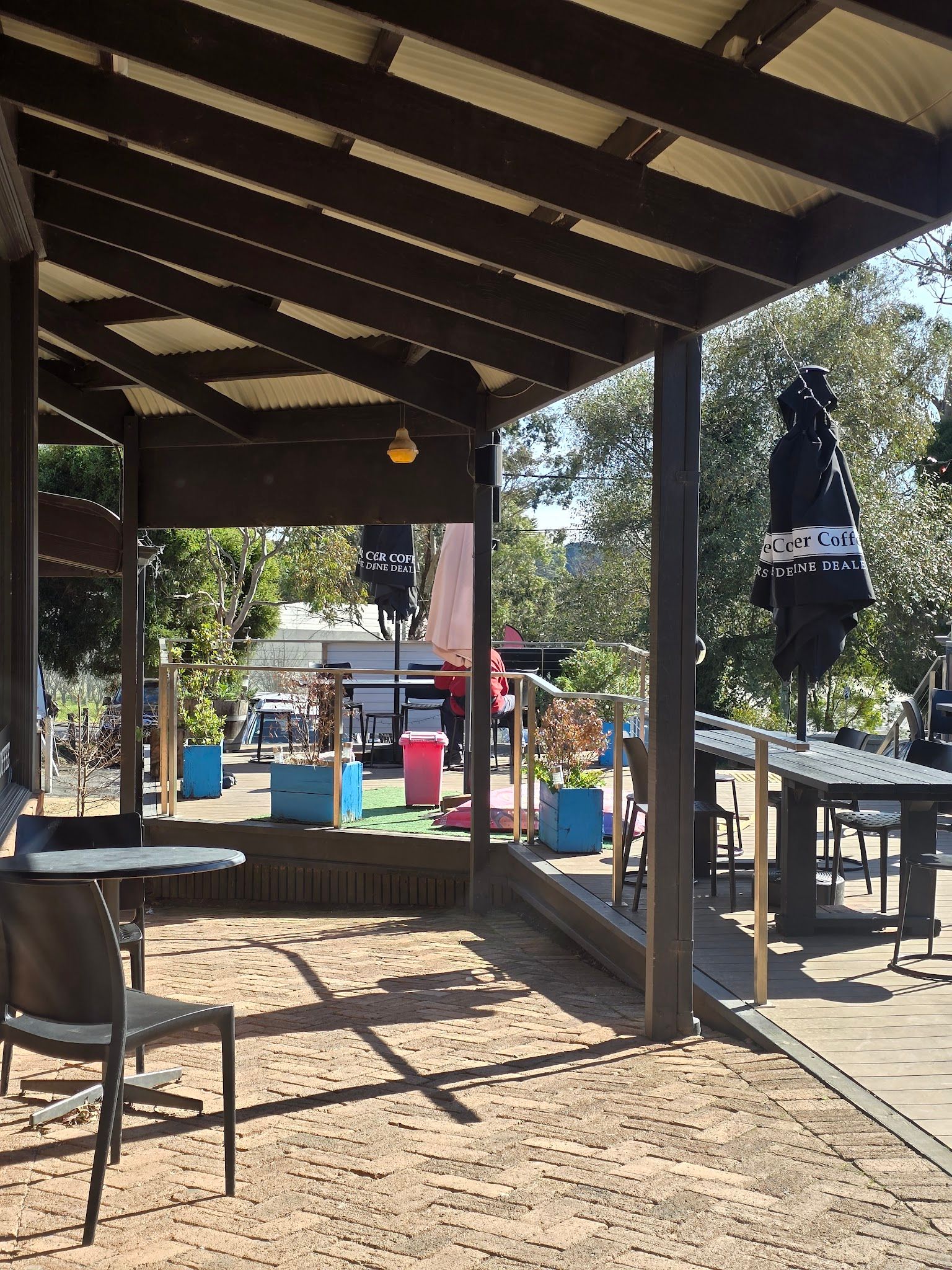 Outdoor alfresco deck at Red Hill Cafe, viewed from under corrugated iron pergola with timber tables and sunny garden beyond