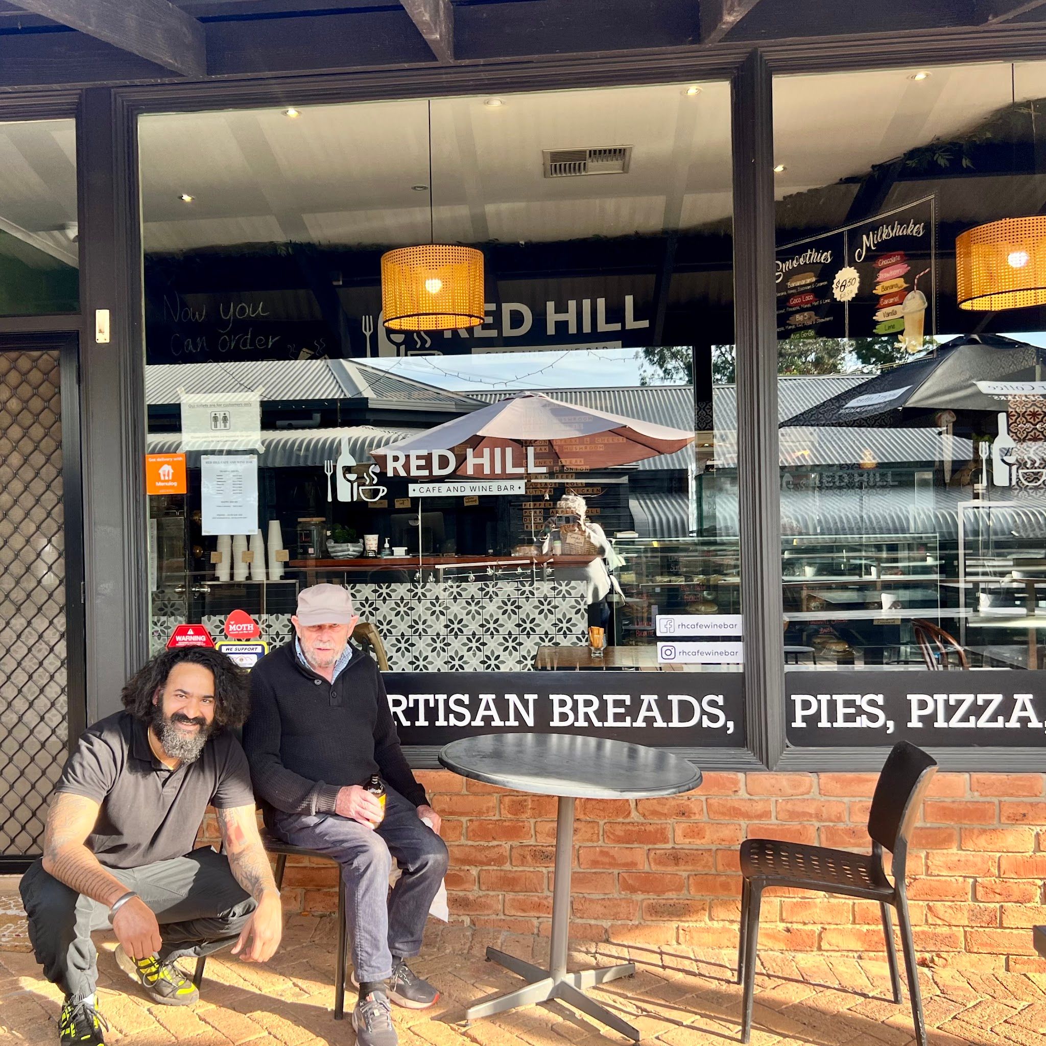 Two men pose outside the Red Hill Cafe and Wine Bar shopfront with glass windows showing the interior