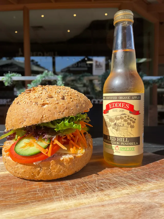 Veggie burger on seeded bun with salad and a bottle of Eddies Red Hill organic apple juice on a timber board