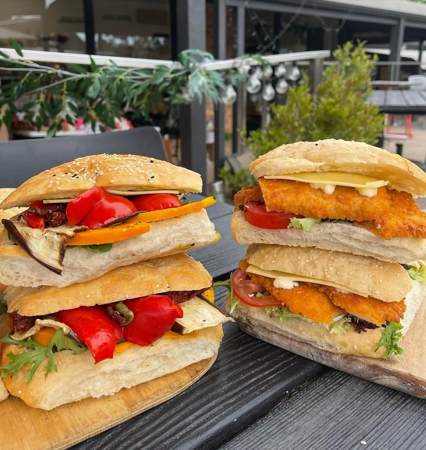 Two large stacked focaccia rolls on wooden boards — one veggie, one crumbed chicken — with outdoor cafe in background