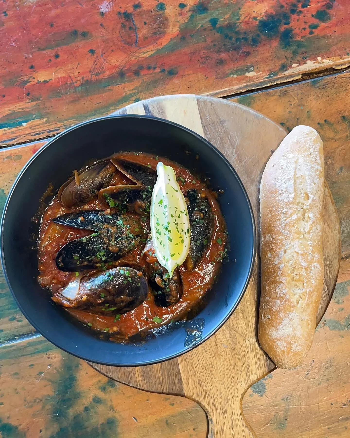 Mussels in tomato broth with a boiled egg and sourdough baguette on a wooden board, vibrant painted surface below