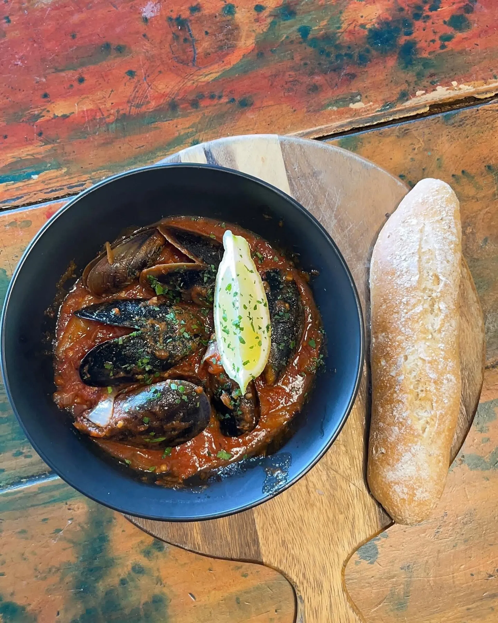 Mussels in tomato broth with a boiled egg and sourdough baguette on a wooden board, vibrant painted surface below
