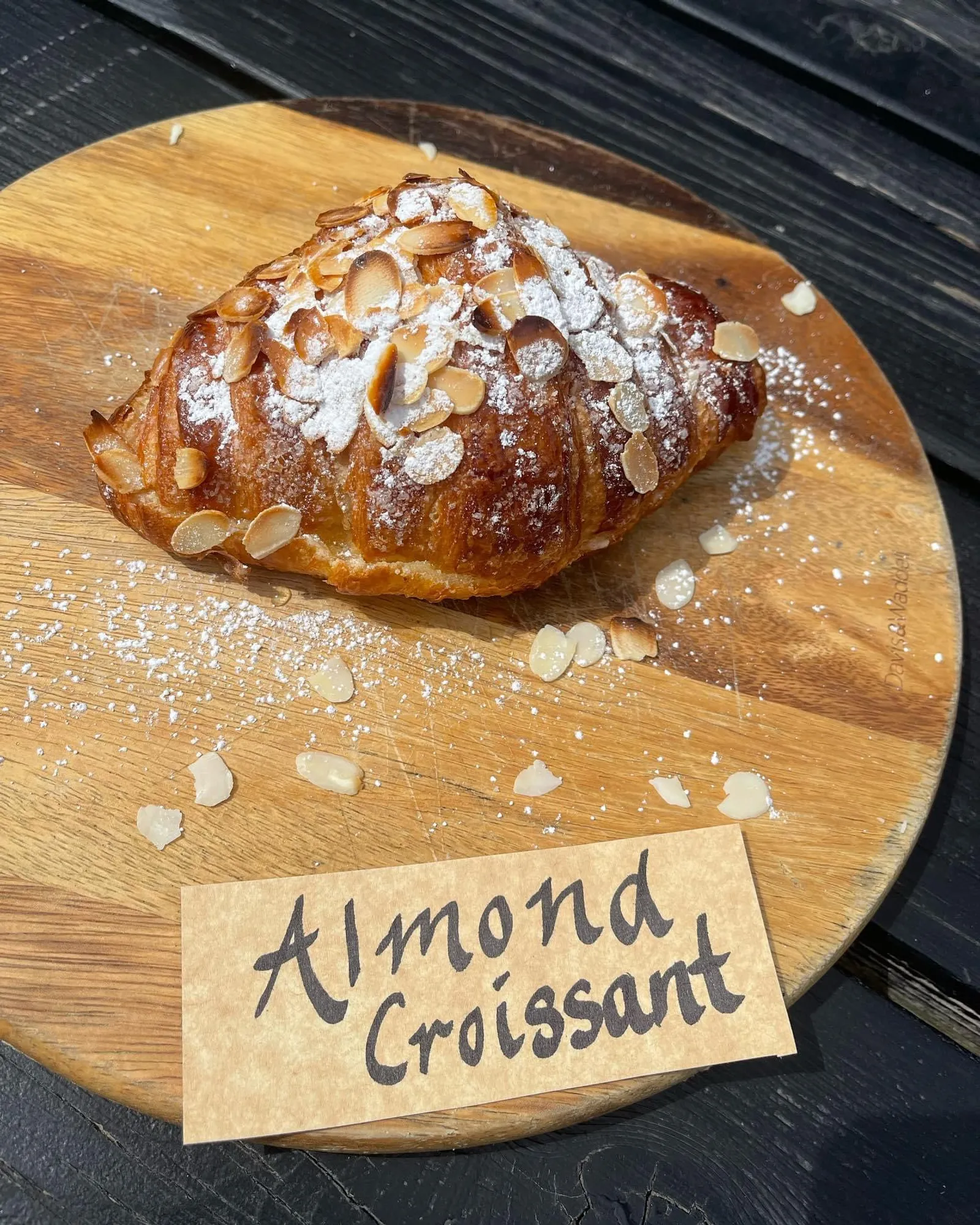 Almond croissant dusted with icing sugar and flaked almonds on a round timber board with handwritten label